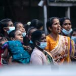 A group of women gather outdoors, wearing masks and colorful clothing, capturing a moment of community and resilience.